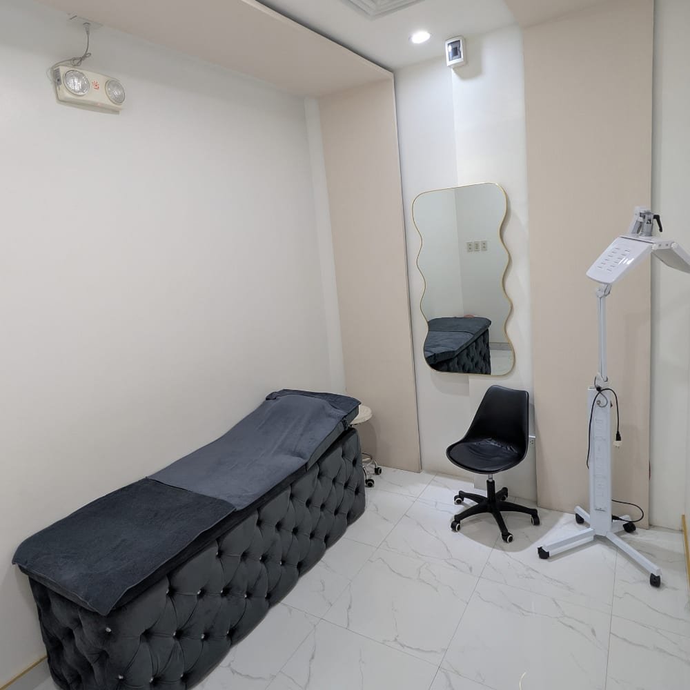 A private treatment room at The Better Face, with a tufted grey treatment bed, a wave-edged mirror and an LED light panel