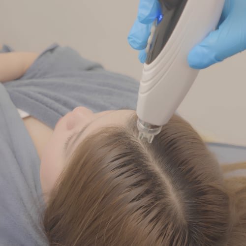A clinician in blue gloves holding a needle-free handheld applicator to a client's parting