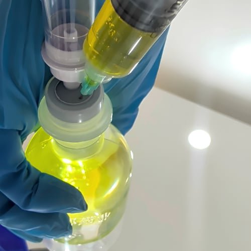 A clinician in blue gloves adding solution from a syringe into a yellow IV bottle